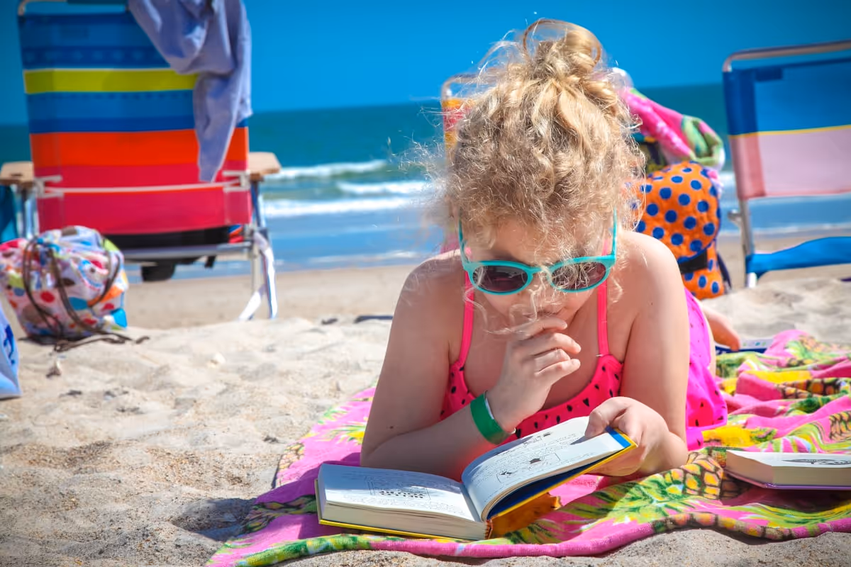 Criança com óculos de sol lendo livro na praia ao sol