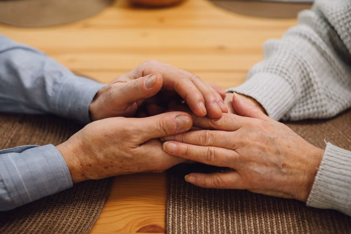 Mãos de um casal de idosos entrelaçadas sobre uma mesa de madeira