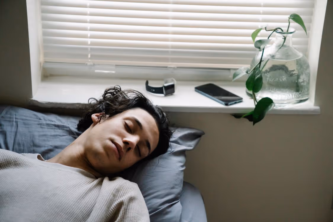 Jovem dormindo em cama com smartwatch e celular sobre a janela ao lado